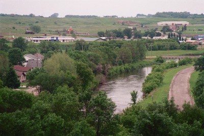 [Small river with shallow banks. A road near the right bank and trees and homes on the left bank.]